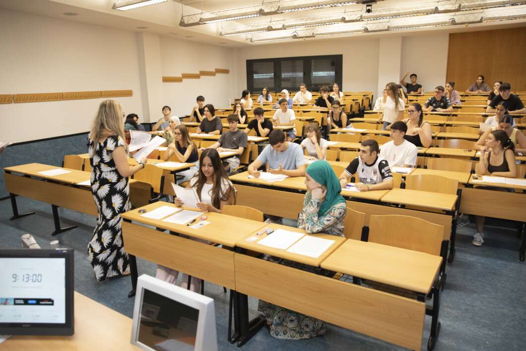 Instructor standing at the front of a large lecture hall, distributing papers to a diverse class of students at tiered wooden desks.