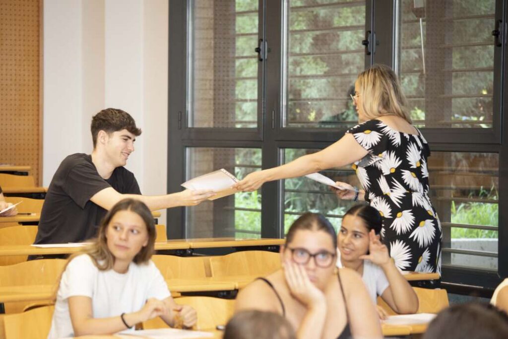 Instructor hands a paper to a male student in a lecture hall as other students look on.