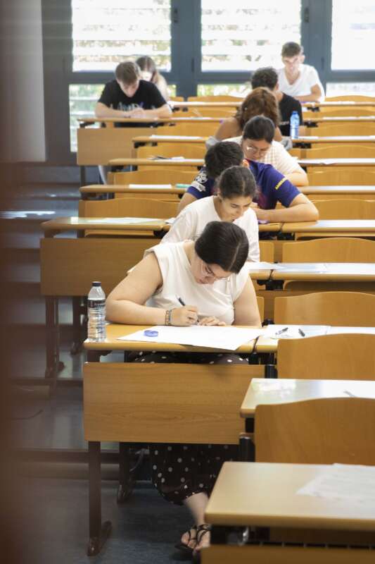 Student writing at a desk in a classroom during an exam; others sit in rows behind.