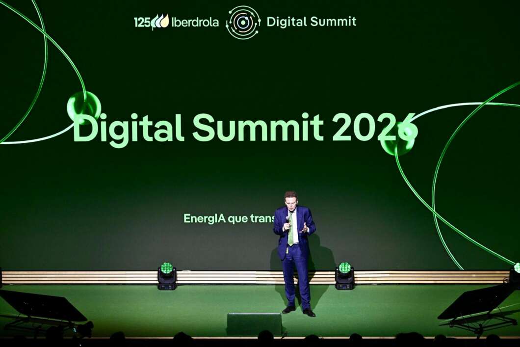 Male presenter in a blue suit stands on a green-lit stage with a large 'Digital Summit 2026' banner behind him, at a corporate summit.