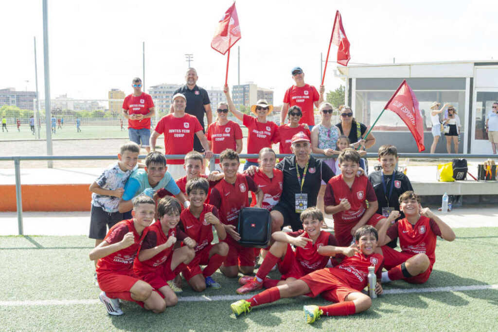 Group of young soccer players in red uniforms posing with coaches on a field, several holding red flags behind them.