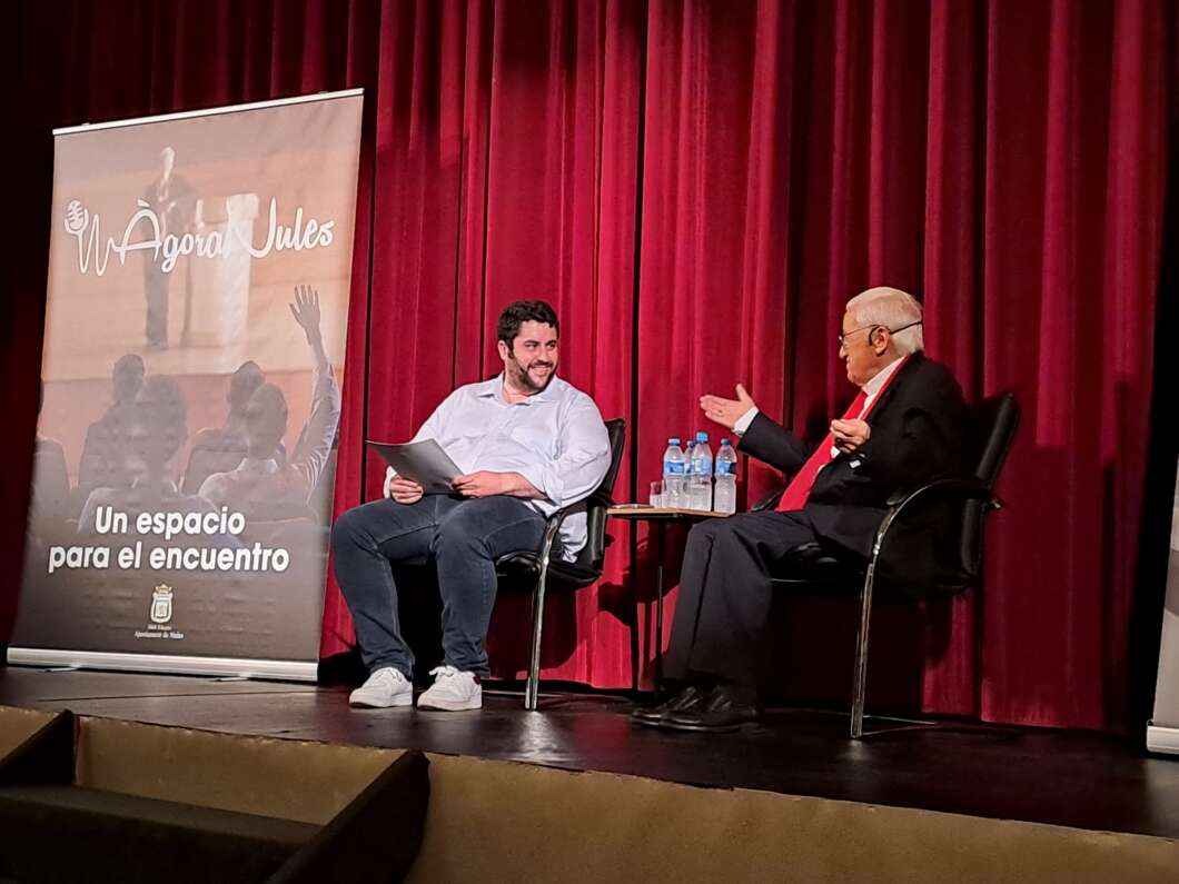 Two men sit in chairs on a stage with red curtains, one holding papers and the other gesturing during a conversation, a banner reads 'Un espacio para el encuentro'.