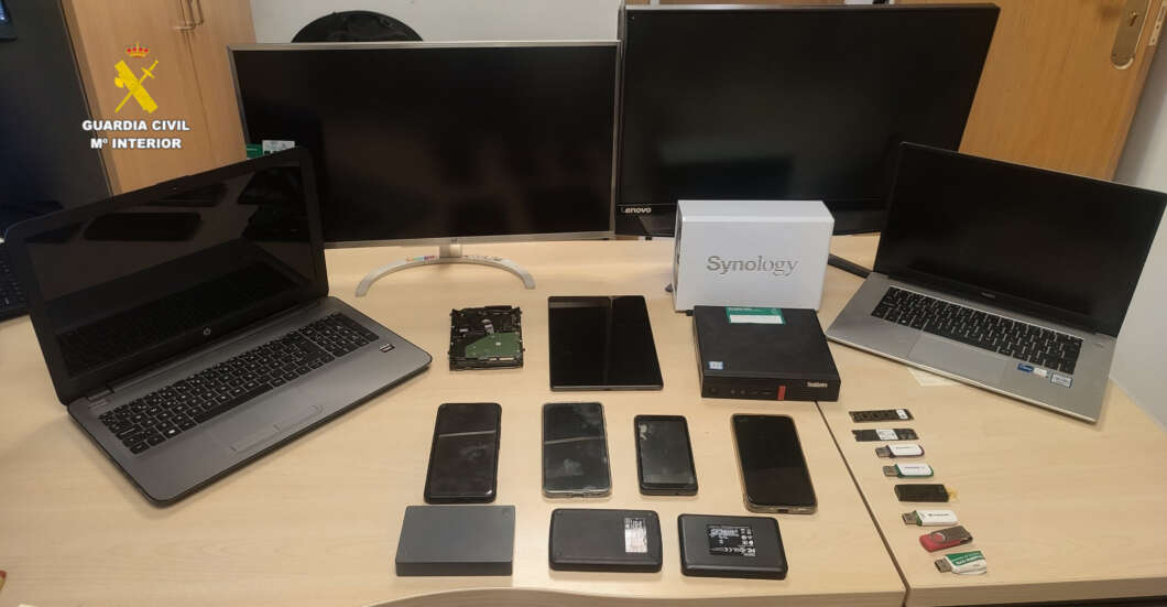 Tech setup on a desk: laptops, a monitor, a Synology NAS box, external hard drive, several smartphones, USB drives and memory cards; Guardia Civil emblem top-left.