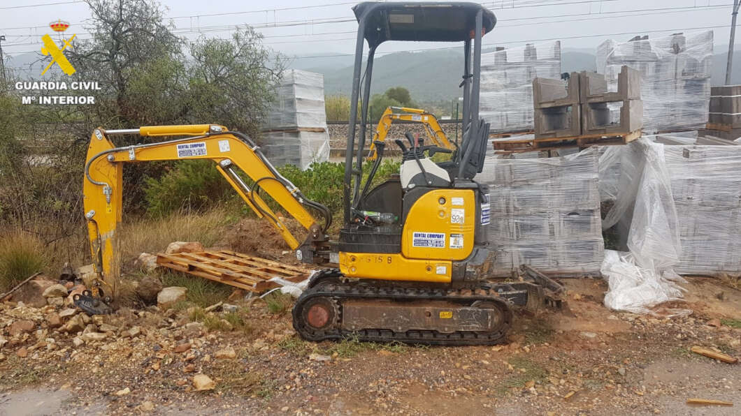 Small yellow excavator on a rocky construction site with pallets wrapped in plastic in the background; Guardia Civil logo in the top left corner.