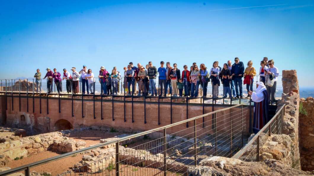 Group of tourists standing along a railing at an ancient ruin site on a sunny day, a guide in traditional dress stands at the right side.