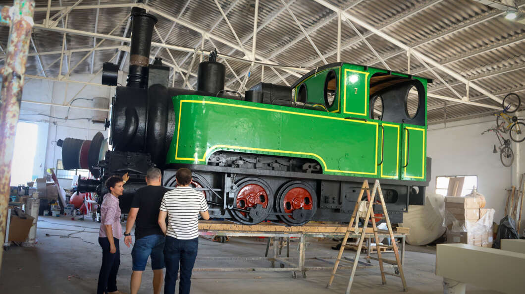 Large green vintage steam locomotive being inspected by four people in a workshop. pieces and tools are visible around.