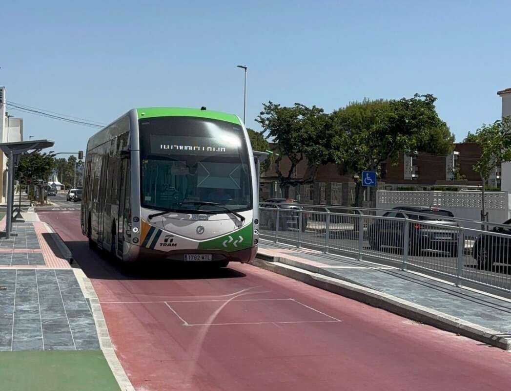 Front view of a green-and-white tram traveling on a red transit lane beside a sidewalk, with trees and parked cars in the background.
