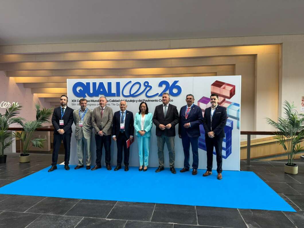 Nine professionals in suits and a turquoise outfit pose on a blue carpet in front of a conference banner reading QUALI... 26.
