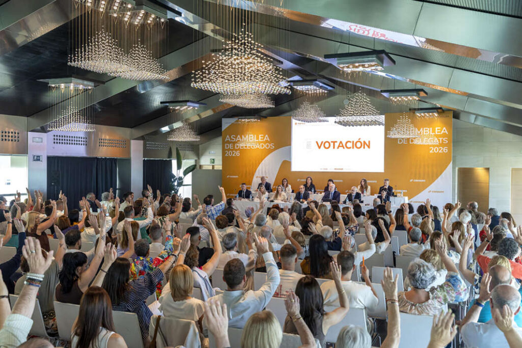 Large conference room with a panel on stage and a screen that reads 'VOTACIÓN' as an audience raises hands to vote.