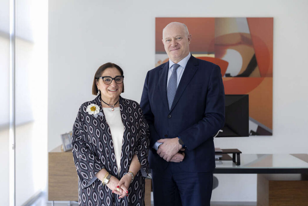 Two professionally dressed people standing in a modern office, smiling at the camera, likely colleagues or clients.