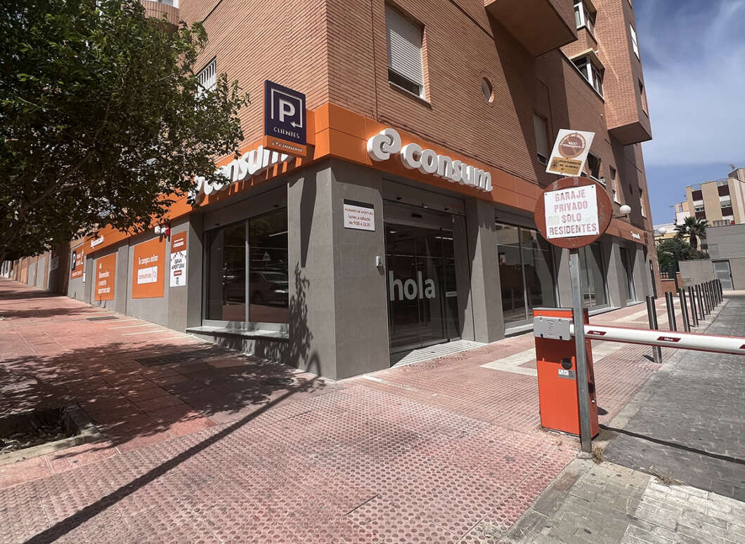 Street corner storefront in a brick building with orange signage reading 'e-consum'; large glass windows show 'hola' inside, and a parking barrier is in the foreground.