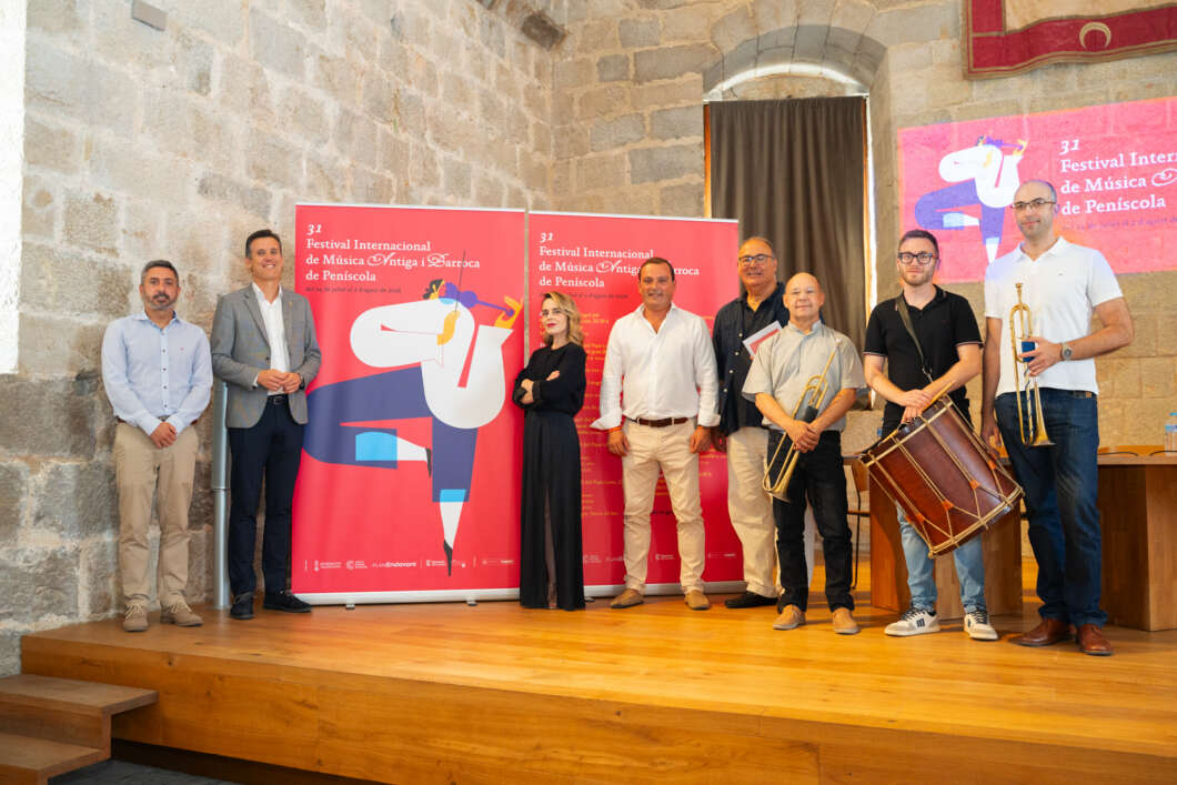 Group of eight people on a wooden stage with brass instruments and a bass drum, posing in a historic venue during a music festival, with red festival banners behind them.