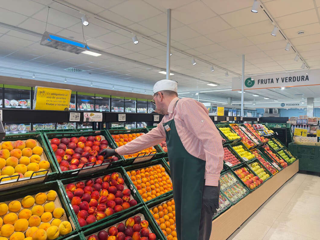 Store worker in a white hat and green apron selects fruit from a display in the supermarket produce section.