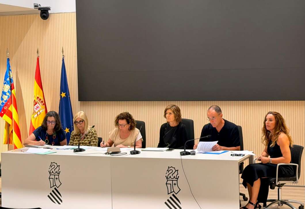 Panel of six people seated at a long white desk with microphones and papers in front, flags (Spain and EU) displayed in a conference room and a large blank screen behind them.