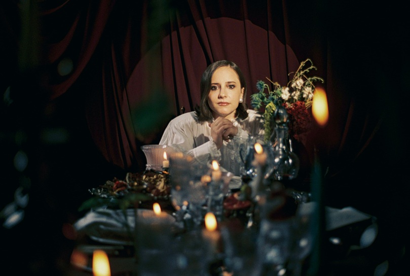 Woman in a white blouse sits at a candlelit dining table with flowers, looking at the camera.