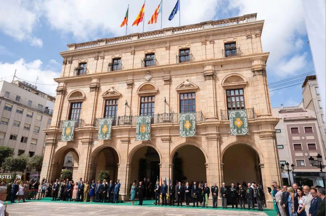 Grand neoclassical building with arches, flags atop, and a ceremonial crowd on a green carpet in front of banners.