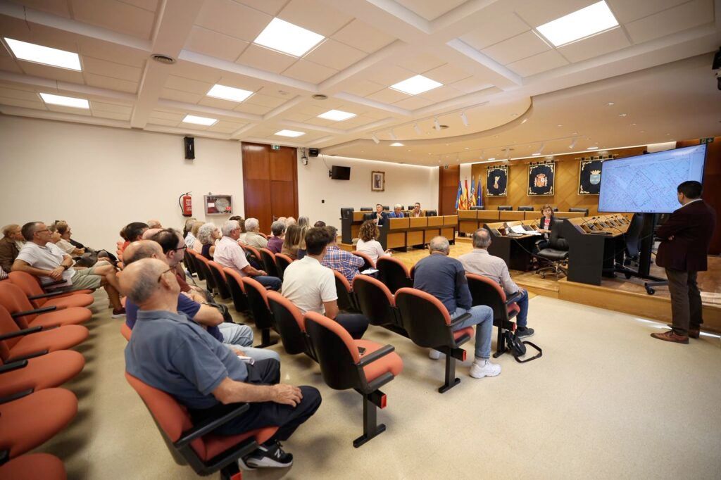 Audience seated in a council chamber listening to a presenter at a podium with a large screen displaying a map.