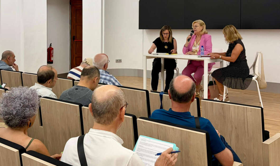 Panel discussion: three women sit at a table at the front, one speaking into a microphone, while an audience watches from wooden seats.