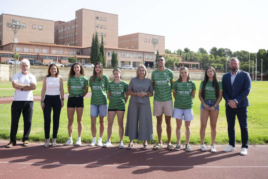 Group of people standing on a running track in front of a brick campus building; several wear green team jerseys with facsa logo.