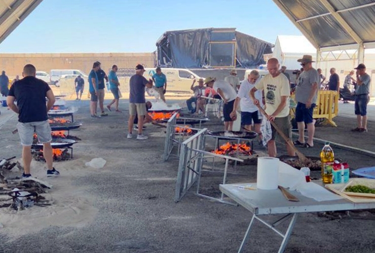 Group of people cooking over multiple open-flame grills under a canopy at an outdoor barbecue event.