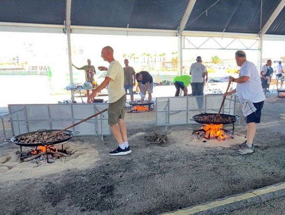 Men grilling over large open flames in circular pans under a canopy at an outdoor barbecue event