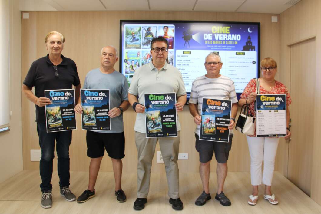 Five adults stand indoors in a row, each holding a poster for a 'Cine de verano' film event in front of a large display screen.