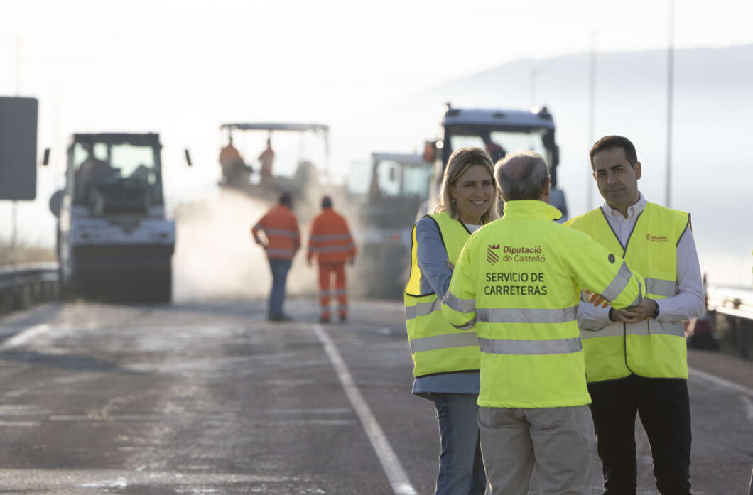 Road workers in high-visibility vests talk on a highway as maintenance trucks operate in the background.