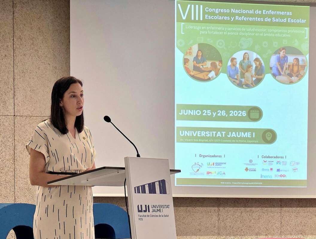 Woman presenting at a podium with a microphone; projection screen shows a green conference poster for the VII Congreso Nacional de Enfermeras Escolares at Universitat Jaume I.