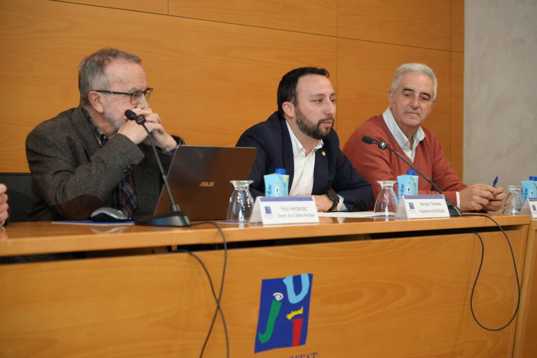 Panel of three men speaking at a wooden conference table with microphones and nameplates in front of them.