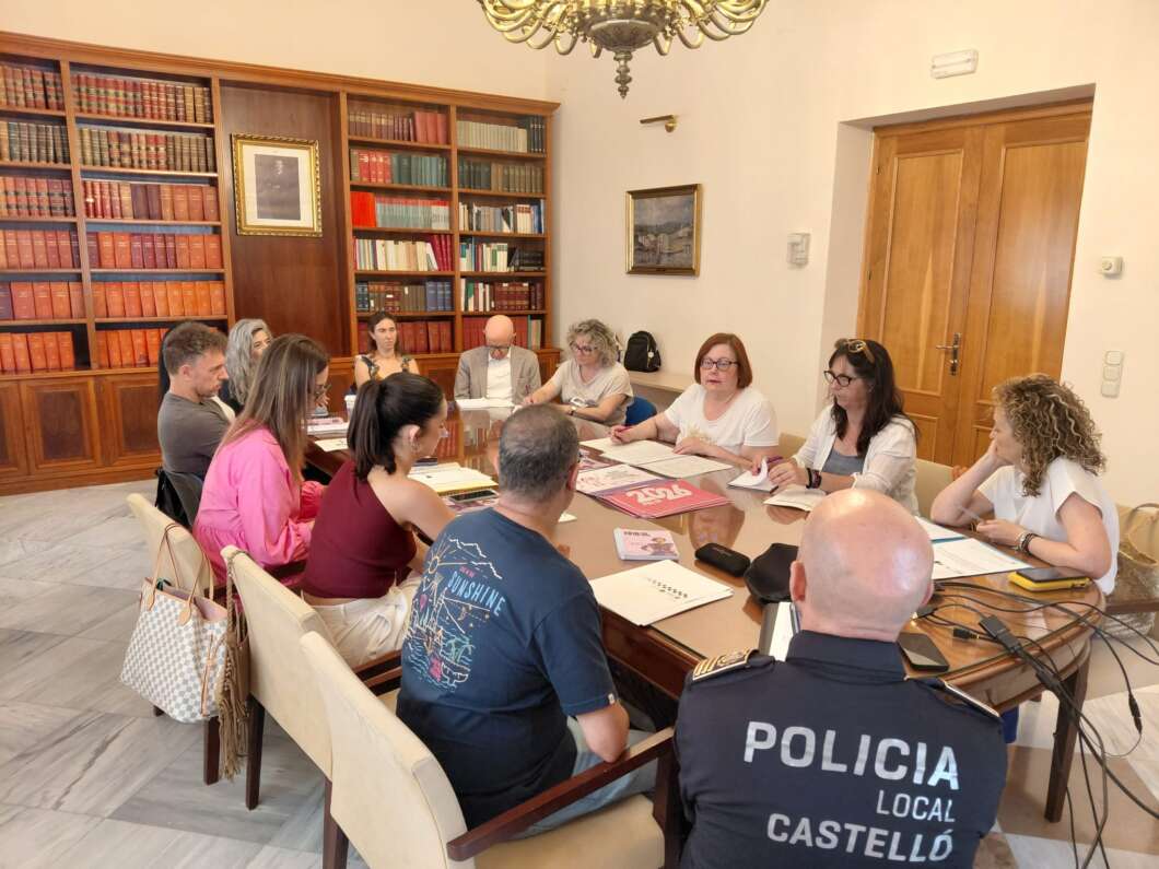 Group of people seated around a large wooden conference table in a library-like room, discussing documents; a local police officer wearing a 'Policia Local Castelló' jacket sits at the foreground left.