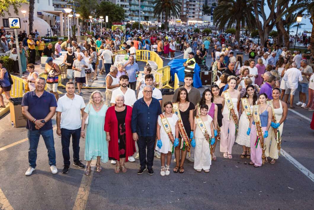 Group of parade participants with sashes and gloves posing for a photo on a crowded waterfront street.