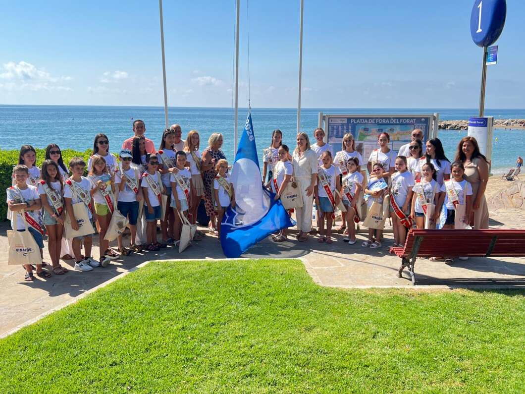 Group of children and adults posing together on a seaside promenade, some wearing sashes and holding a blue flag near a map board by the beach.