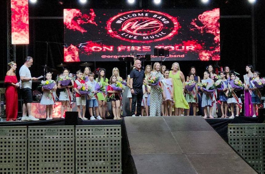 Group of children and adults on a stage holding bouquets during a concert awards event, with a red-lit backdrop reading 'On Fire Tour' and 'Welcome Band'.