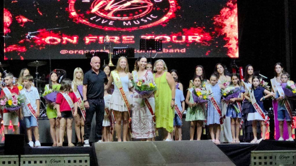 Children and adults on a stage after a pageant, some wearing sashes and holding flowers, with a bright screen behind them.