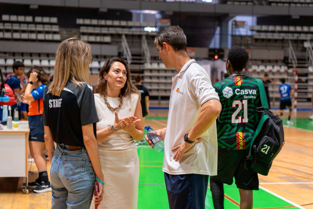 Staff member talks with two adults on a gym court; the woman in white gestures as the man in a white polo holds a water bottle nearby on the sports floor.