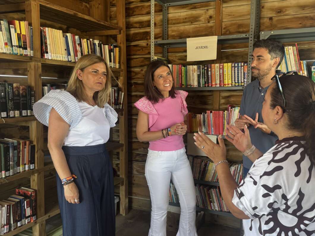 Four adults stand in a rustic wooden library, chatting in front of shelves of colorful books; a 'Juvenil' sign is visible.