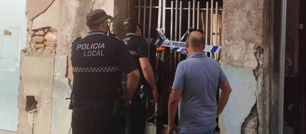 Local police block access to a building with blue-and-white tape across a barred doorway outside a damaged wall in daylight.