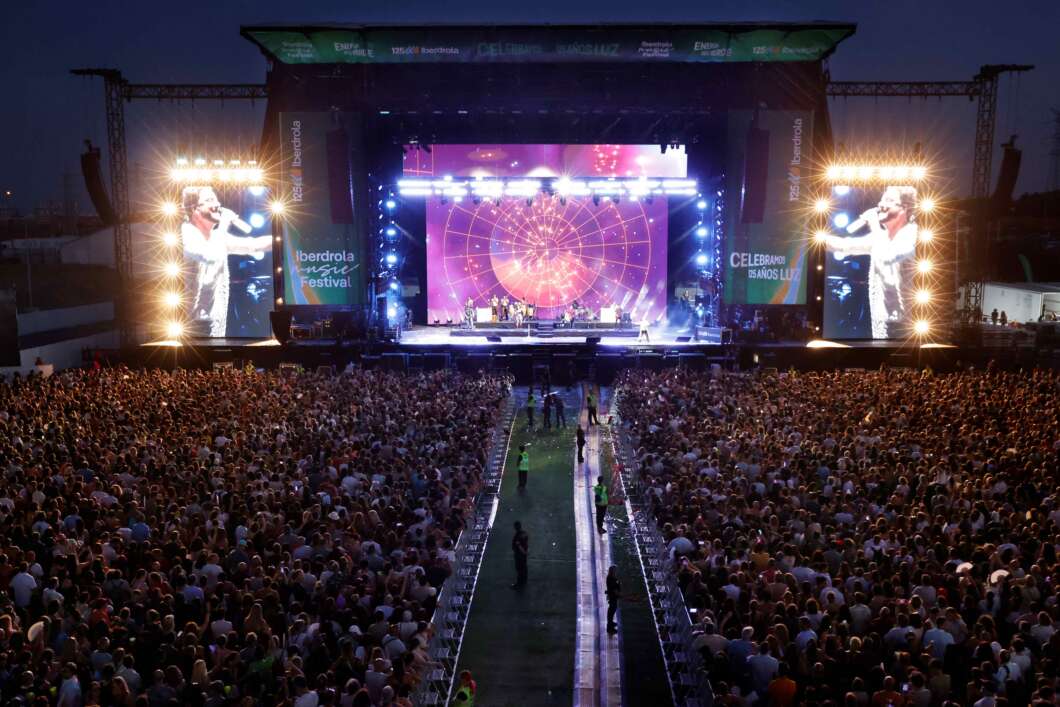 Large outdoor concert stage at night with bright lights and two tall side screens overlooking a packed audience.