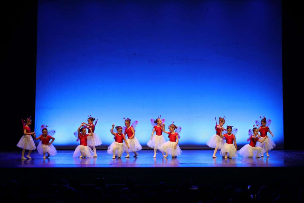 Group of young dancers in red leotards and white tutus performing on a blue-lit stage in a theater.
