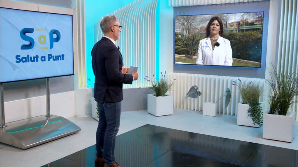 Male TV host stands in a bright studio, facing a large screen showing a female doctor in a white coat on a video call.
