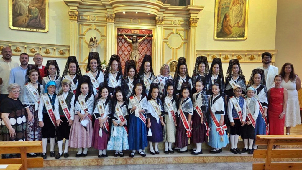 Group of girls in traditional dresses and sashes posing on church steps in front of an ornate altar and crucifix.