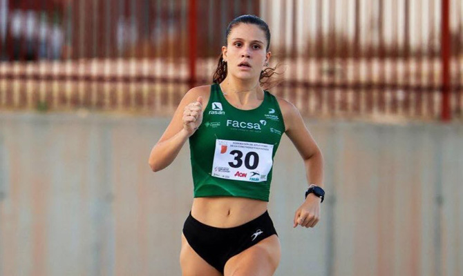 Female long-distance runner in a green race top with bib 30, mid-race on a track edge.