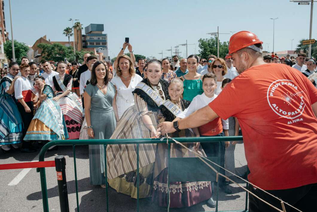 Crowd of people in traditional dresses watches a man in a red shirt and hard hat handling ropes at a street festival barrier.