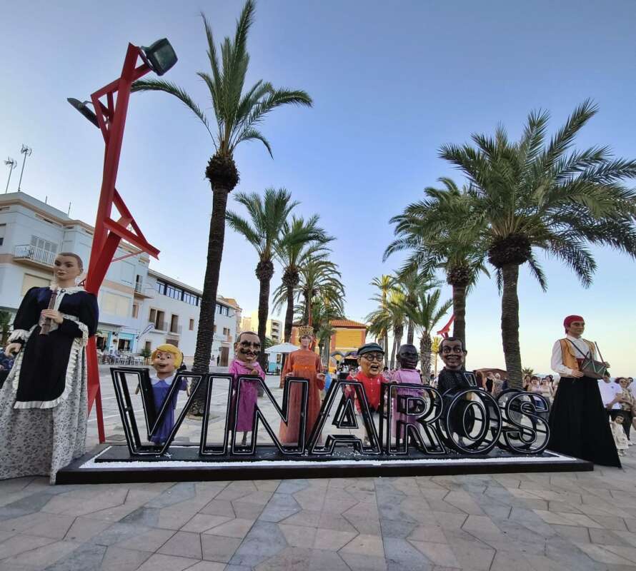 Outdoor festival scene with oversized puppets and a large 'VINAROS' sign on a palm-lined promenade.
