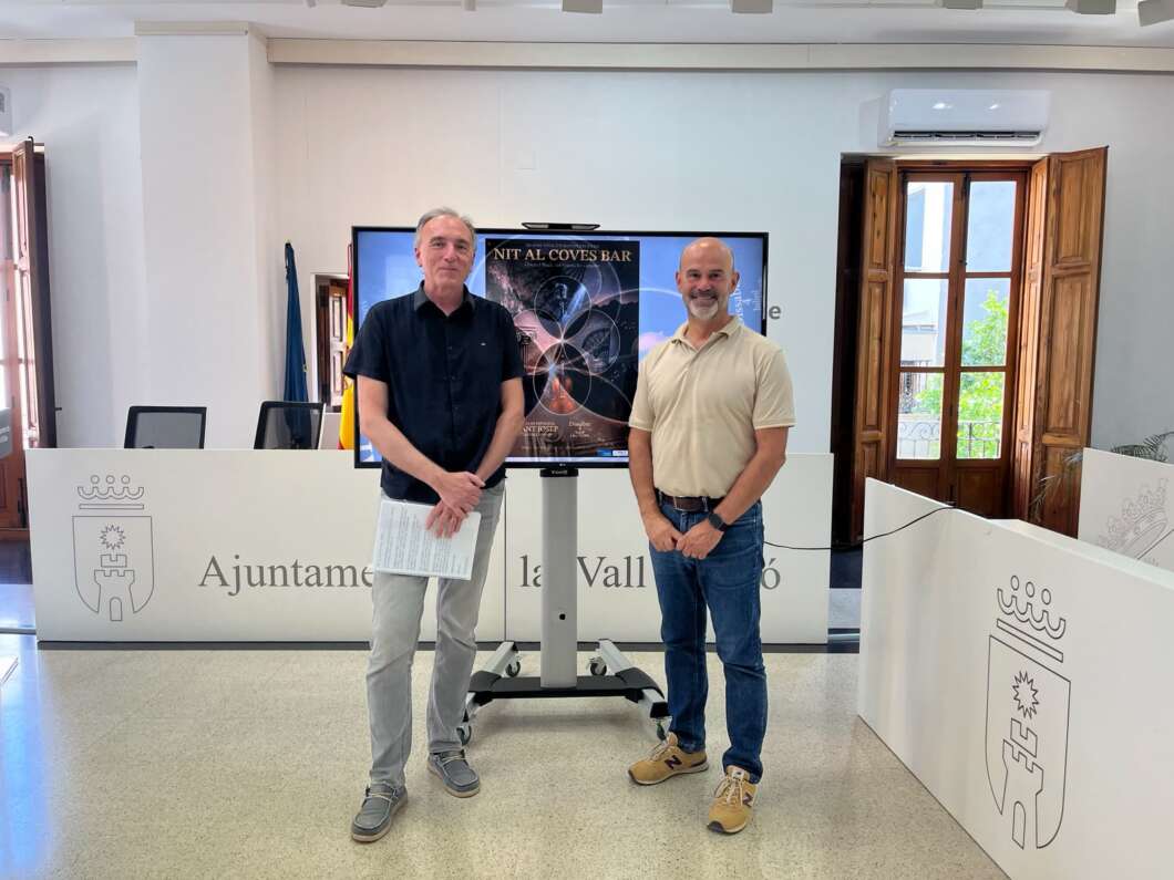 Two men stand in a municipal office in front of a large screen displaying a poster behind them.
