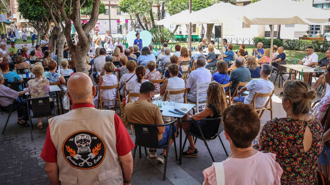 Crowd seated at an outdoor community event in a sunny plaza, with a speaker on a stage under umbrellas.