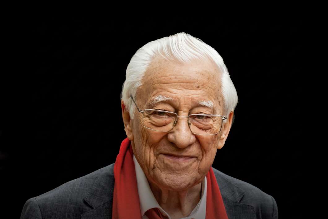 Portrait of an elderly man with white hair and glasses, wearing a dark suit and red scarf, smiling softly against a black background.