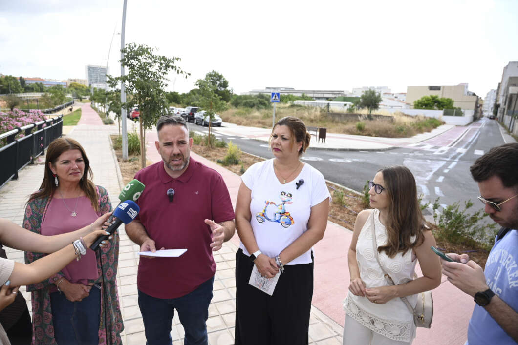 Five people stand on a sidewalk during an outdoor interview; a man in a maroon polo speaks into multiple microphones while others listen nearby.