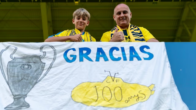 Two sports fans in yellow shirts and scarves hold a banner reading 'GRACIAS 100 anys' with a trophy illustration in a stadium stand.