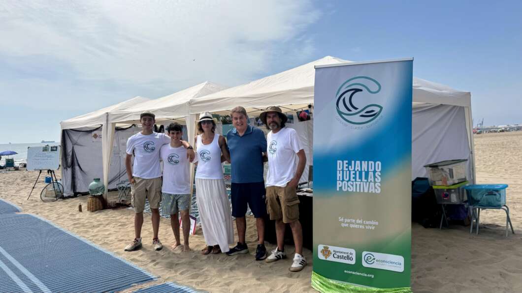 Group of six people standing on a sandy beach beside white tents and a tall banner reading 'Dejando Huellas Positivas' on a sunny day, likely at an outreach event.